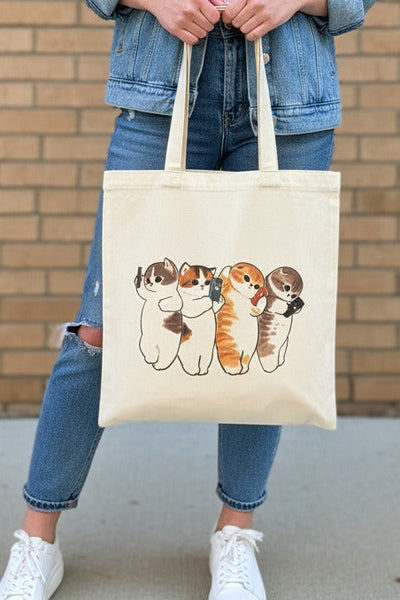 A cream canvas tote bag hanging on a display stand in a retail store, featuring a printed illustration of four cute calico cats holding phones.