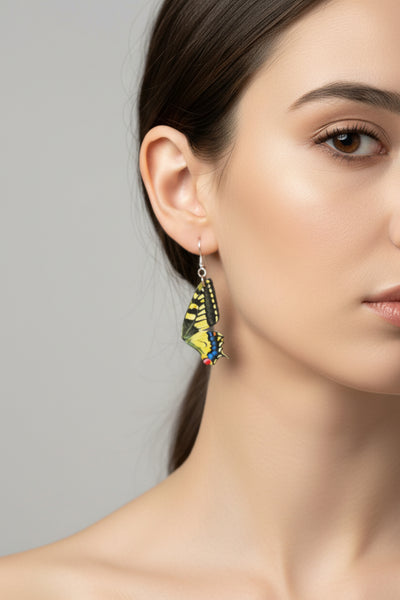 A pair of butterfly-shaped dangle earrings with yellow, black, blue, and red wing details, hanging from silver fishhook wires on a dark gray background.