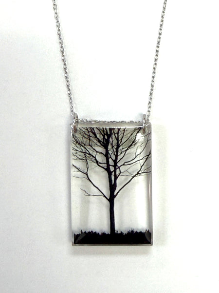 A rectangular pendant necklace featuring a black tree silhouette inside a clear resin or glass panel, suspended from a silver-toned chain against a plain white background.