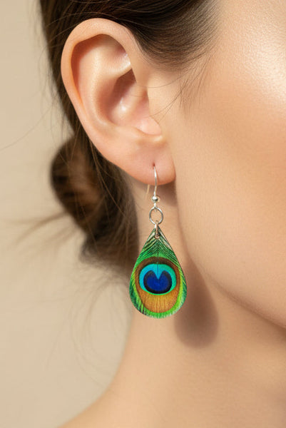 A pair of dangling earrings shaped like peacock feathers, resting on the edge of a closed laptop on a dark textured surface.