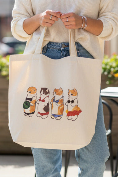 A cream canvas tote bag with four cartoon cat illustrations in colorful outfits, hanging in a retail store.